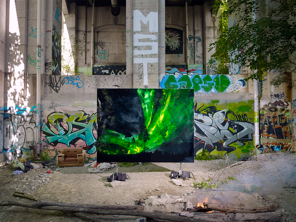 A photograph of a painting within an urban landscape. Graffiti tags cover a cement structure. Tree on right side with shadow of same tree on opposite side. Small fire in foreground. Painting in middle ground of photograph depicting night sky with green northern lights and a tree in centre.