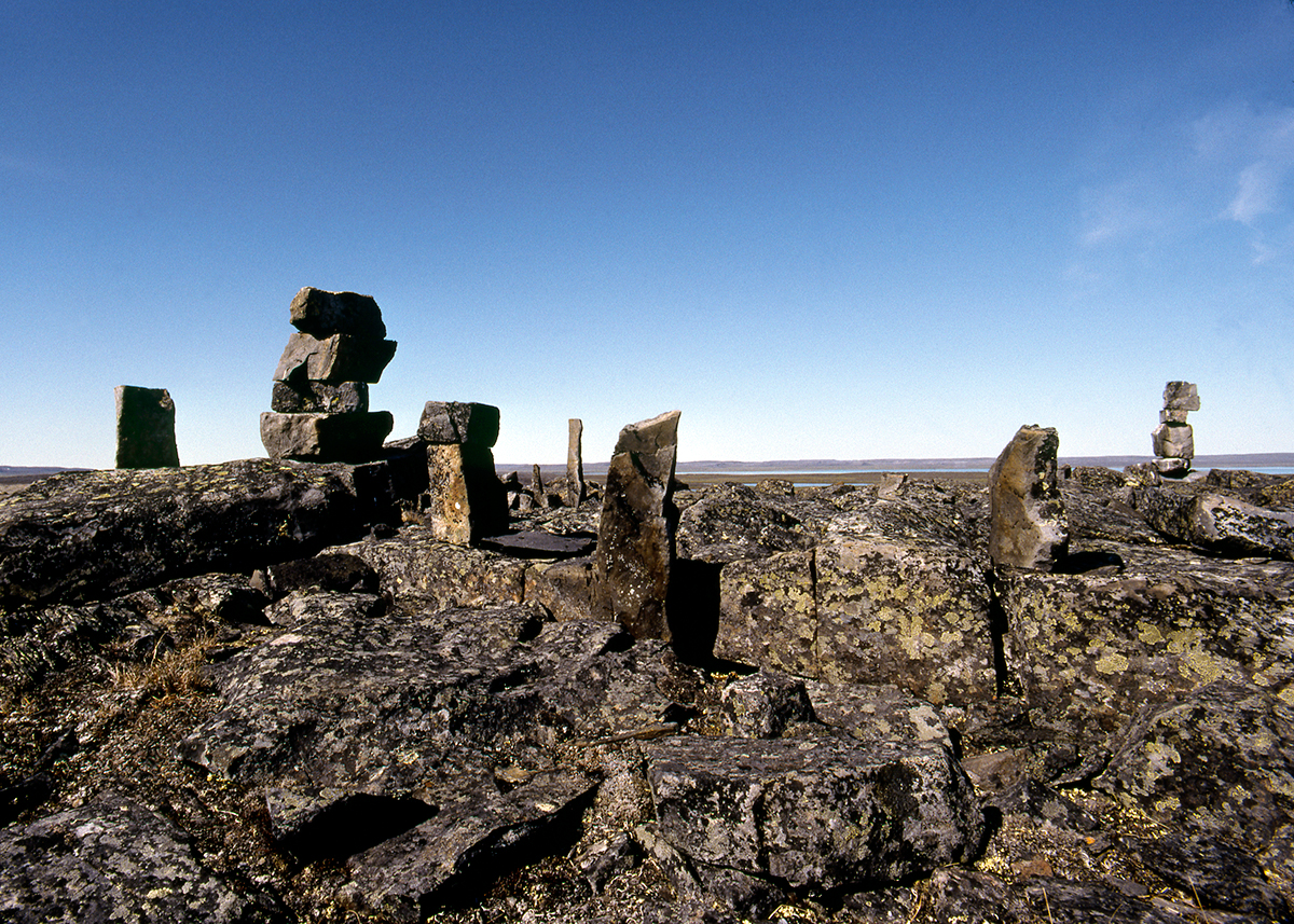 Photograph of rocky landscape in bottom half with eight or more upright stones interspersed across the image in foreground and distance. Lichen growing on the grey rocks. Sliver of pale blue water in far distance with rocky shore on horizon and blue sky above in top half of photograph.