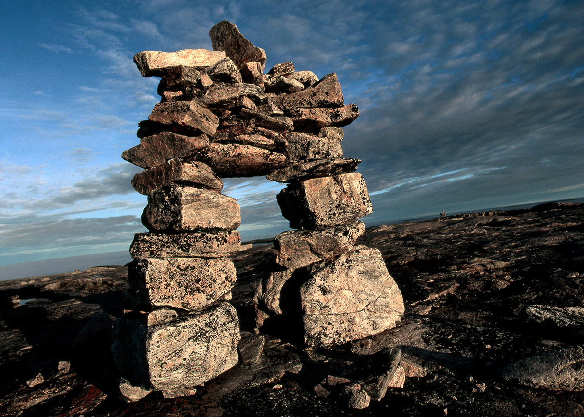 Photograph of rocky landscape with rocks piled in the shape of a doorway. Blue sky above with grey and white clouds.
