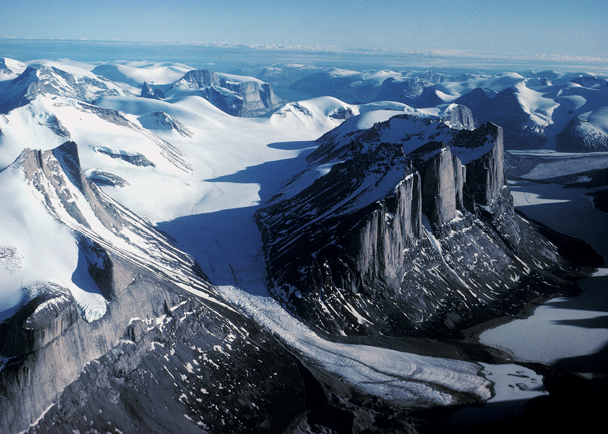 Aerial photograph of snow-covered mountains with body of water in the far distance and blue sky above.