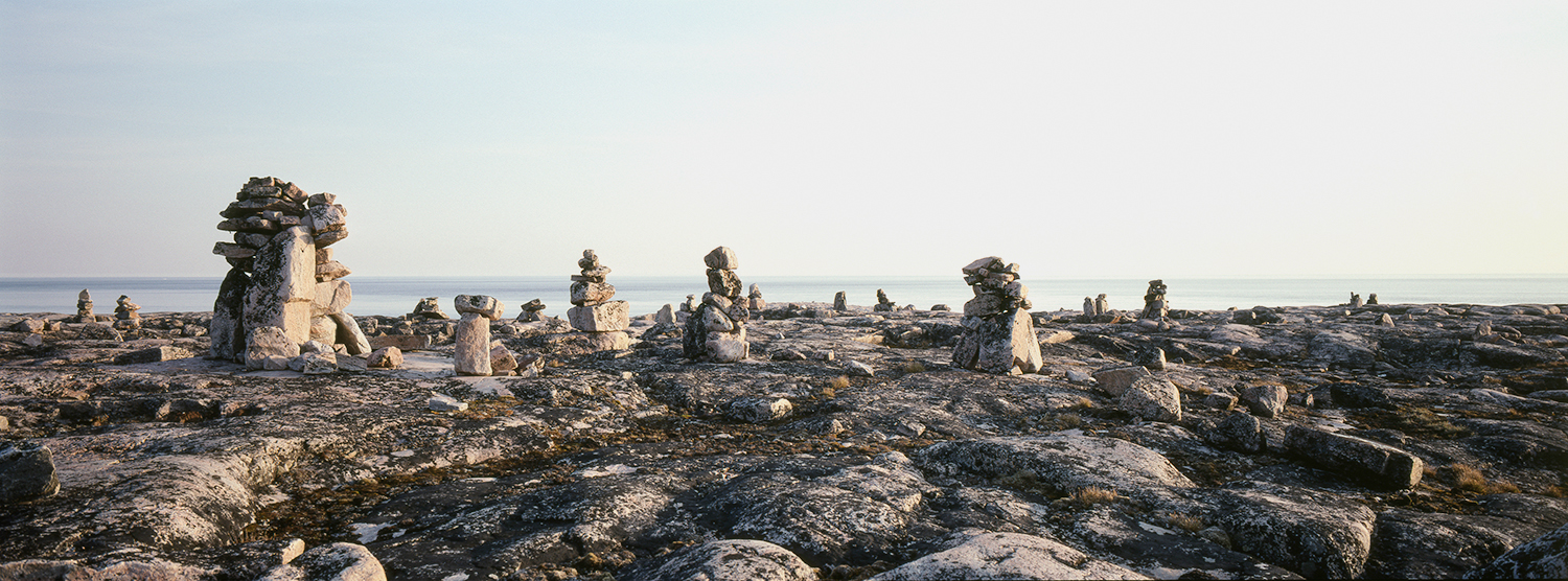 Photograph of rocky landscape with body of water beyond and the sky above. Many piles of stones in various sizes are on the grey rocky land. Moss and lichens are growing on the rocks.