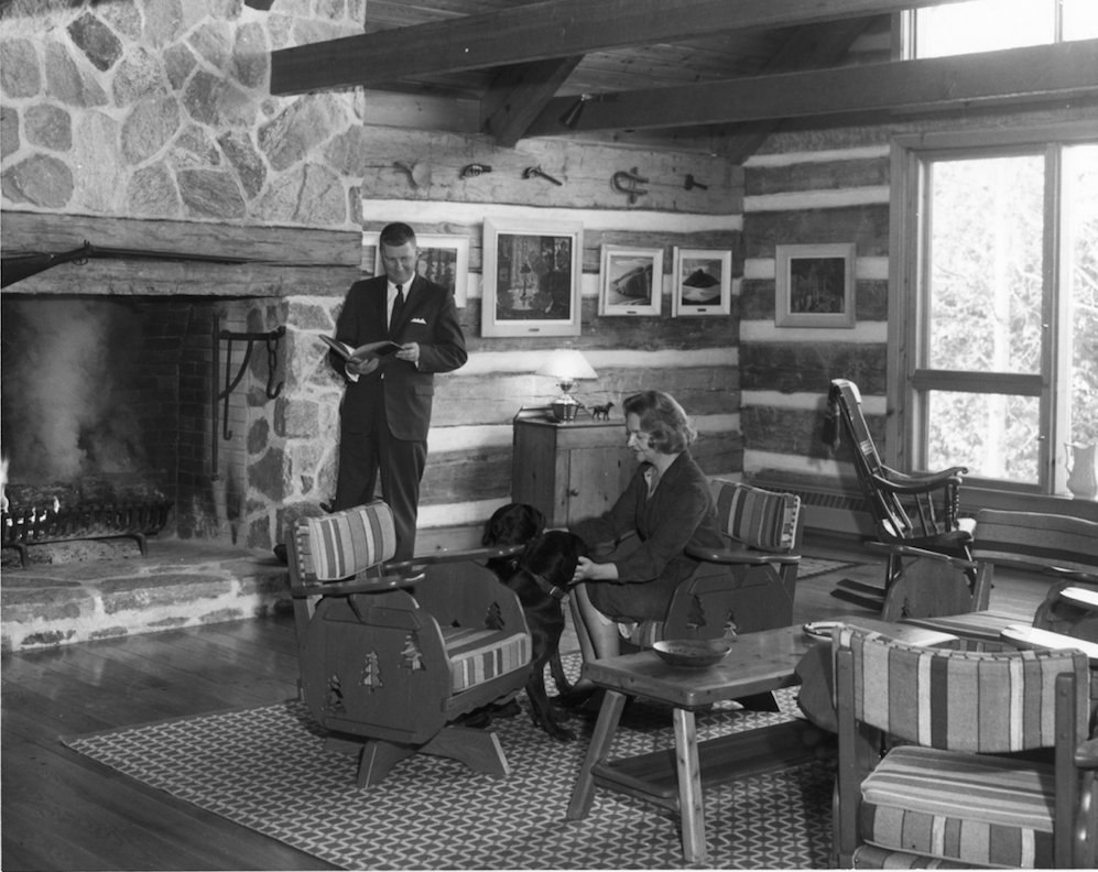 Robert and Signe McMichael Photograph in black and white of a man and woman in a sitting room.The man is standing by an open stone fireplace reading a book. There is a fire in the hearth. The woman is seated on one of a number of chairs. She is petting a black dog. There are five paintings behind them on log and plaster walls and a window with view of trees to the right.