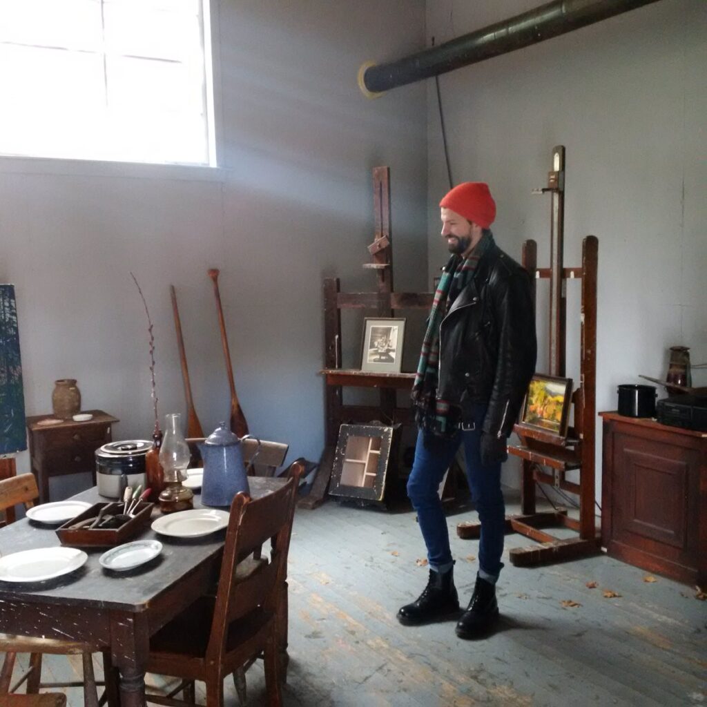 Zachari Logan in Tom Thomson’s shack photo of man wearing a red hat in an artist's studio. Light from upper window streaming in